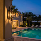 Seen at dusk, this Clérachic wall lantern is lit on a pillar beside a pool, with a villa and palms in the background.