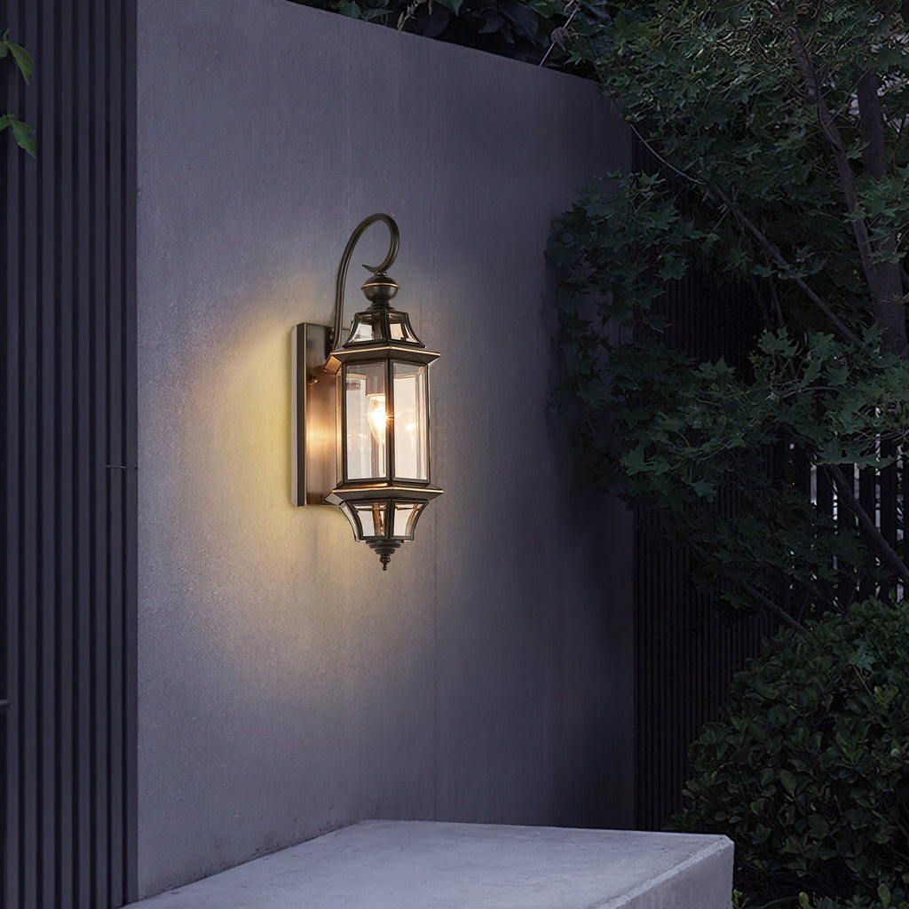 A lit Clérachic wall lantern on a grey garden wall, with dark leaves hanging over from one side.