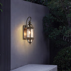 A lit Clérachic wall lantern on a grey garden wall, with dark leaves hanging over from one side.