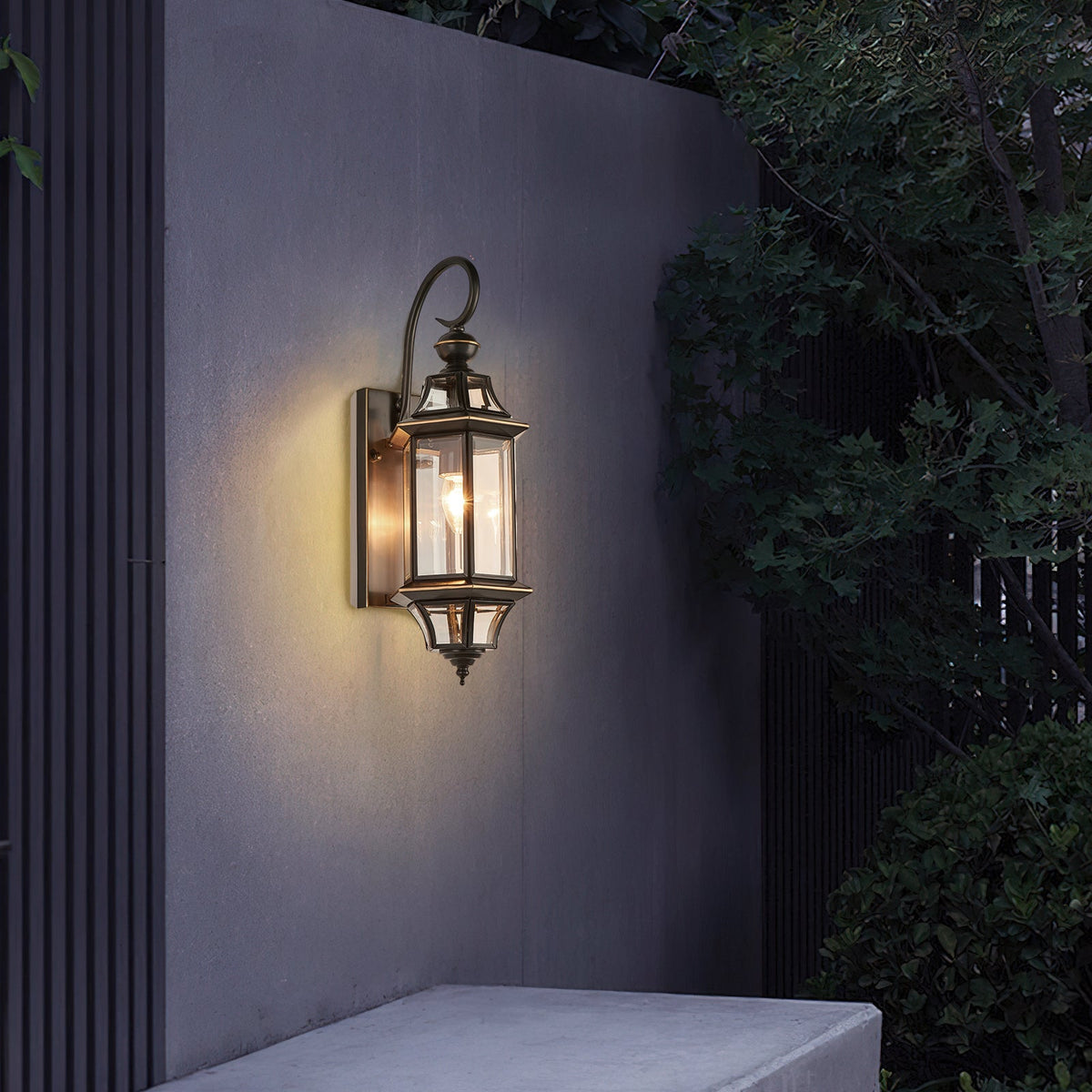 A lit Clérachic wall lantern on a grey garden wall, with dark leaves hanging over from one side.