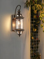 This Clérachic wall lantern sits on a pale wall beside a trellis with climbing leaves, giving off warm light.