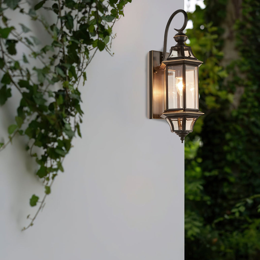 A Clérachic outdoor wall lantern lit against a white wall, with trailing ivy and dark foliage beside it.