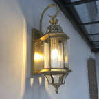 A brass-toned version of this Clérachic wall lantern, lit under a glass canopy, with the curved bracket arm over the top.