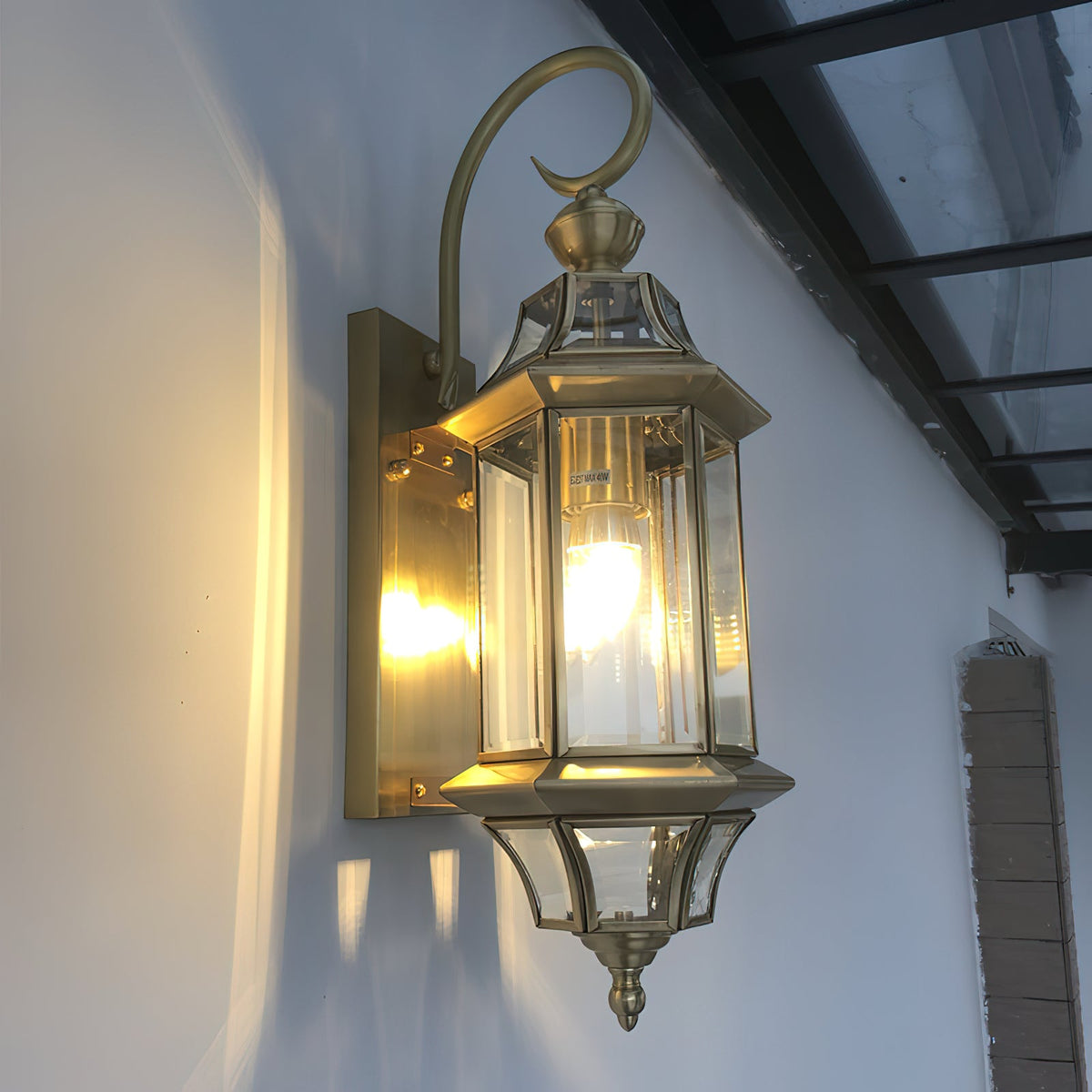 A brass-toned version of this Clérachic wall lantern, lit under a glass canopy, with the curved bracket arm over the top.