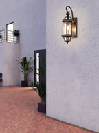 A Clérachic wall lantern on a lilac-grey rendered wall in a courtyard with brick paving and potted plants.