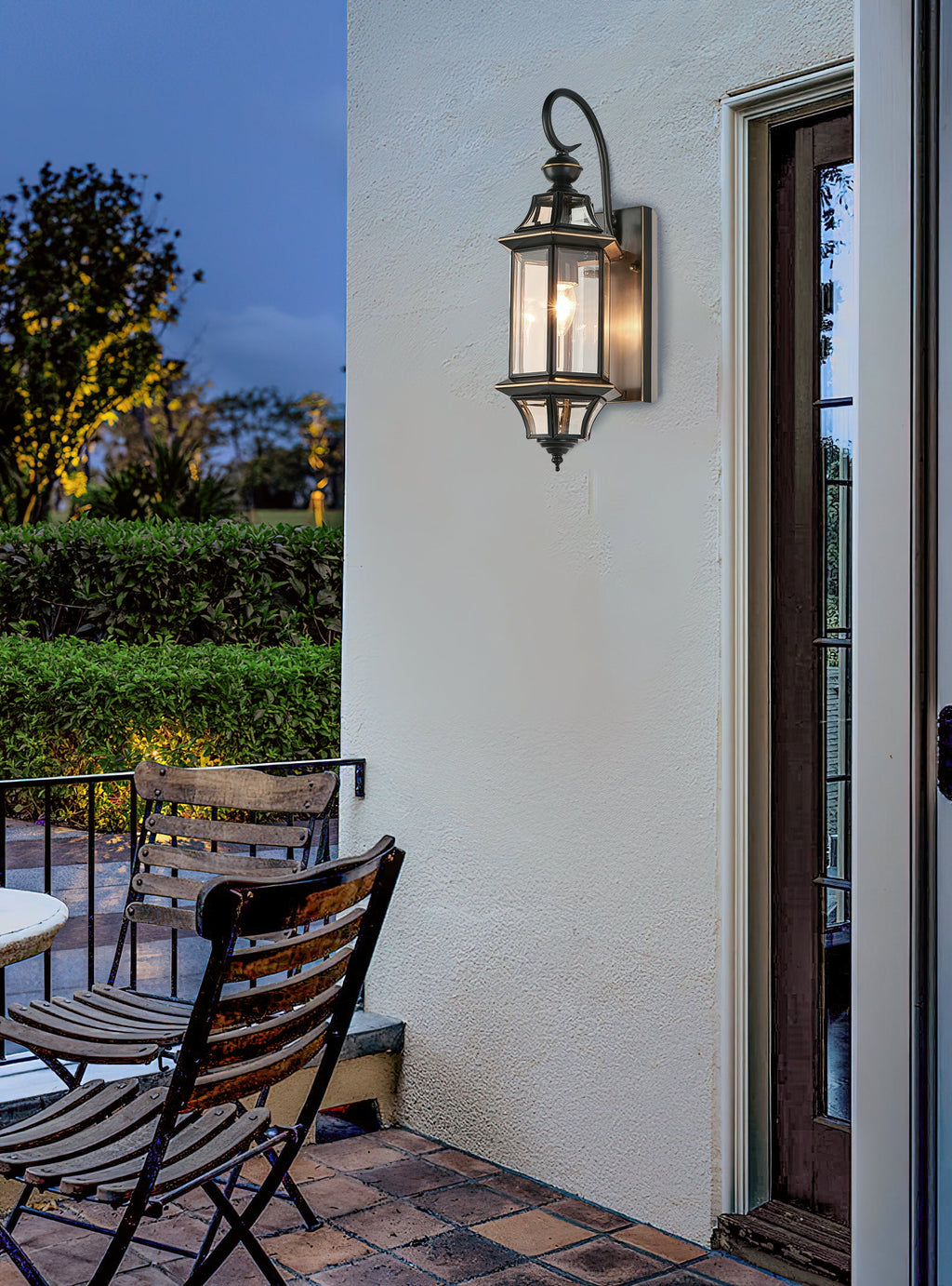 This Clérachic wall lantern hangs on a white wall by a glazed door, with garden chairs and a clipped hedge at dusk.