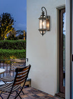 This Clérachic wall lantern hangs on a white wall by a glazed door, with garden chairs and a clipped hedge at dusk.