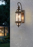 A Clérachic outdoor wall lantern lit on a textured grey render wall, with a softly blurred garden behind.