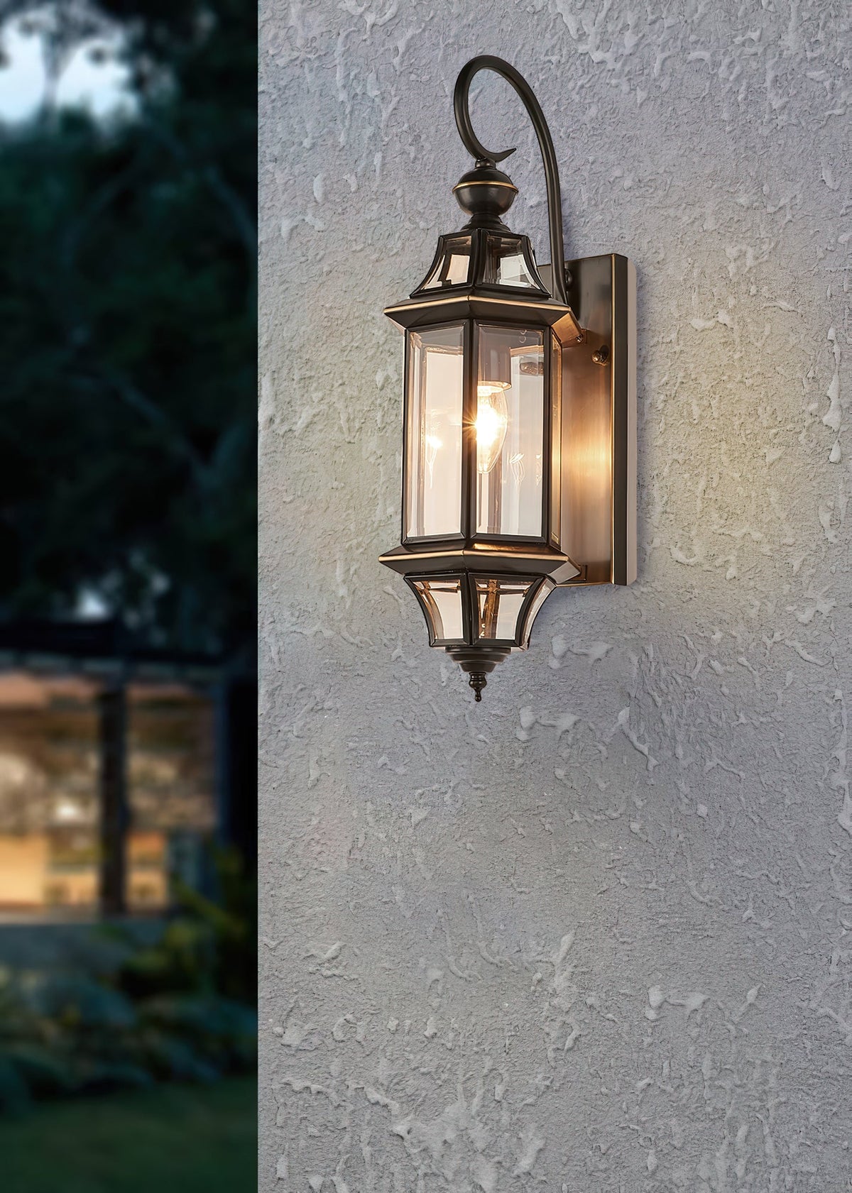 A Clérachic outdoor wall lantern lit on a textured grey render wall, with a softly blurred garden behind.