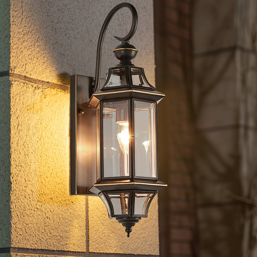 Close view of the curved black arm, clear glass panes and lit bulb of this Clérachic outdoor wall lantern on a stone wall.