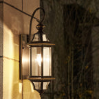 Close view of this Clérachic wall lantern lit at night, its glass panels and black frame against a stone wall.