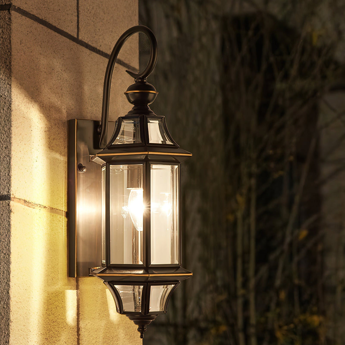 Close view of this Clérachic wall lantern lit at night, its glass panels and black frame against a stone wall.