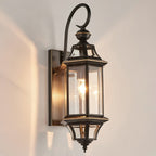 A Clérachic outdoor wall lantern lit on a pale wall, its black frame with thin brass-coloured trim around clear glass.