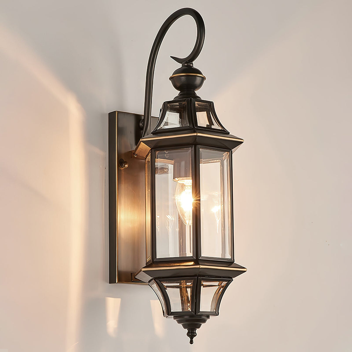 A Clérachic outdoor wall lantern lit on a pale wall, its black frame with thin brass-coloured trim around clear glass.