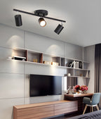 A black Clérachic three-head track light above a living room with a wall-mounted TV, open shelves and a wooden unit.