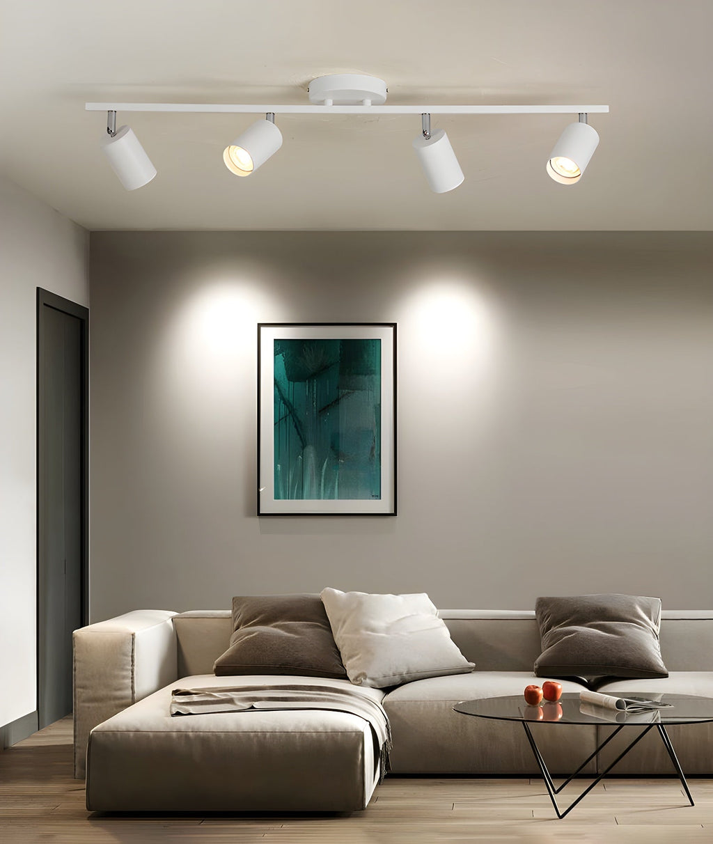 A white Clérachic four-head track light above a lounge with a corner sofa, a glass coffee table and a teal print.