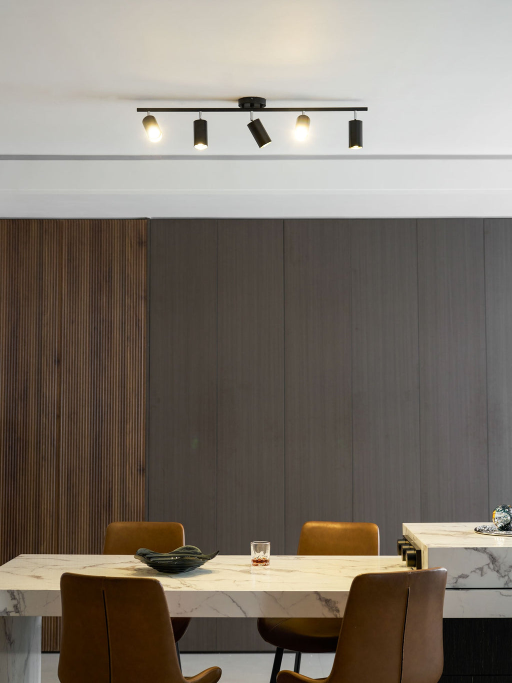 Black Clérachic five-head track light on a white ceiling above a marble table and tan chairs, in a room panelled in grey.