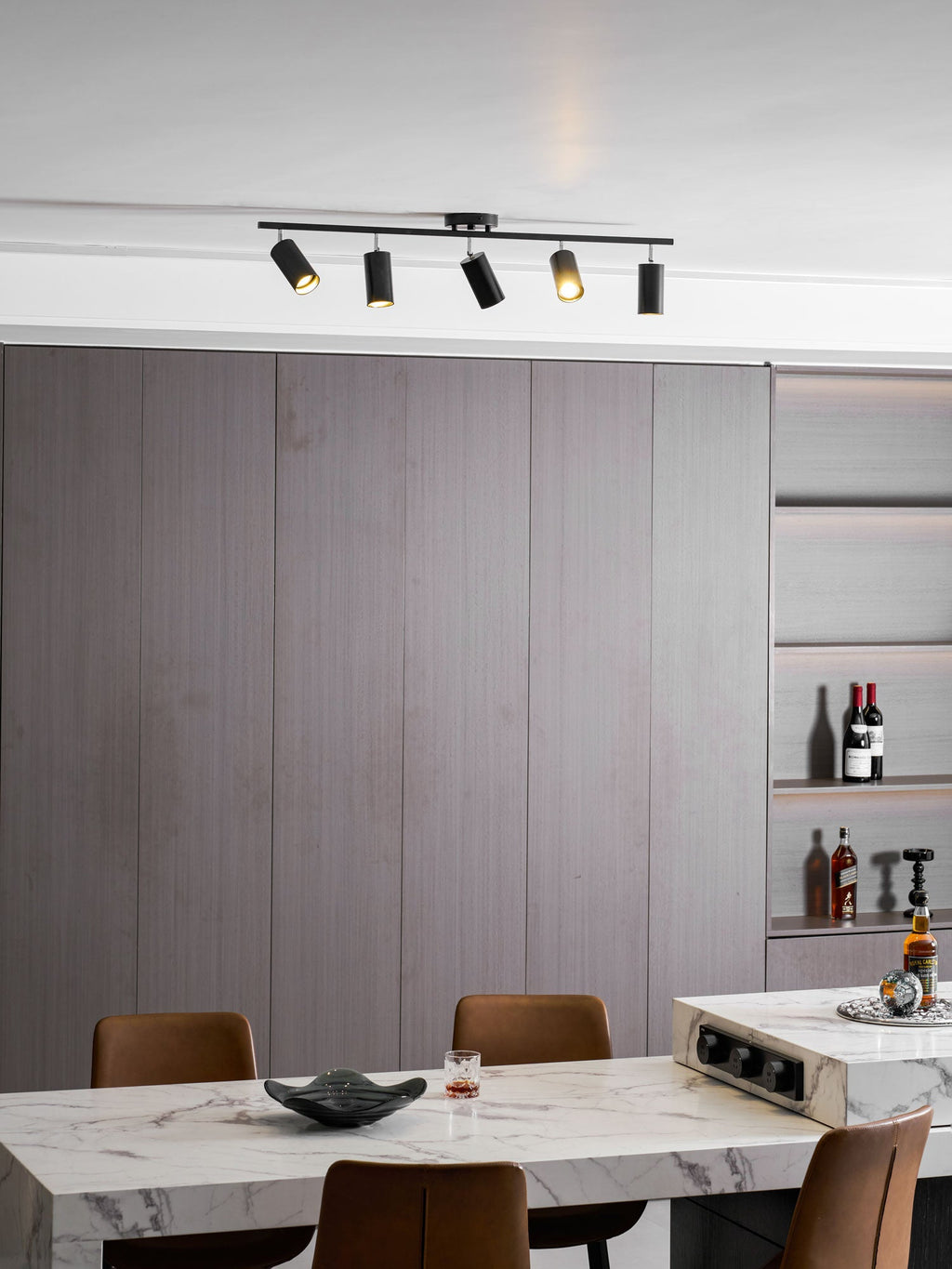 Black five-head track light by Clérachic over a dining room with a marble table, tan chairs and a lit wine shelf.