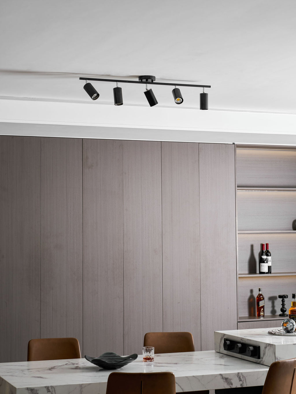 Angled heads on a black Clérachic five-head track light on a white ceiling above grey wall panels and a marble table with a bowl.