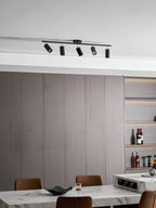 Angled heads on a black Clérachic five-head track light on a white ceiling above grey wall panels and a marble table with a bowl.