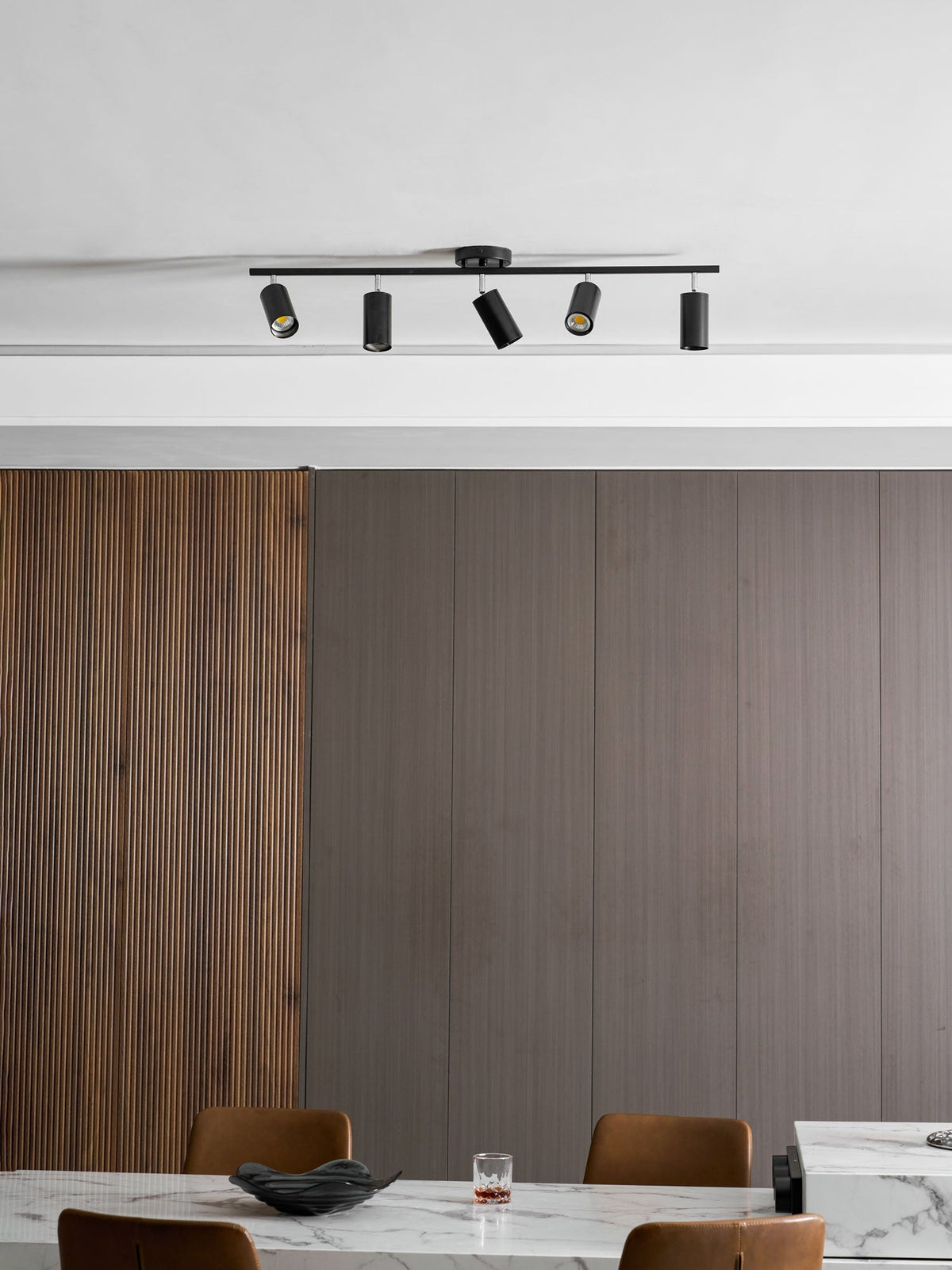 Black Clérachic track light with five heads on a ceiling above ribbed wood panelling and a marble table with tan chairs.