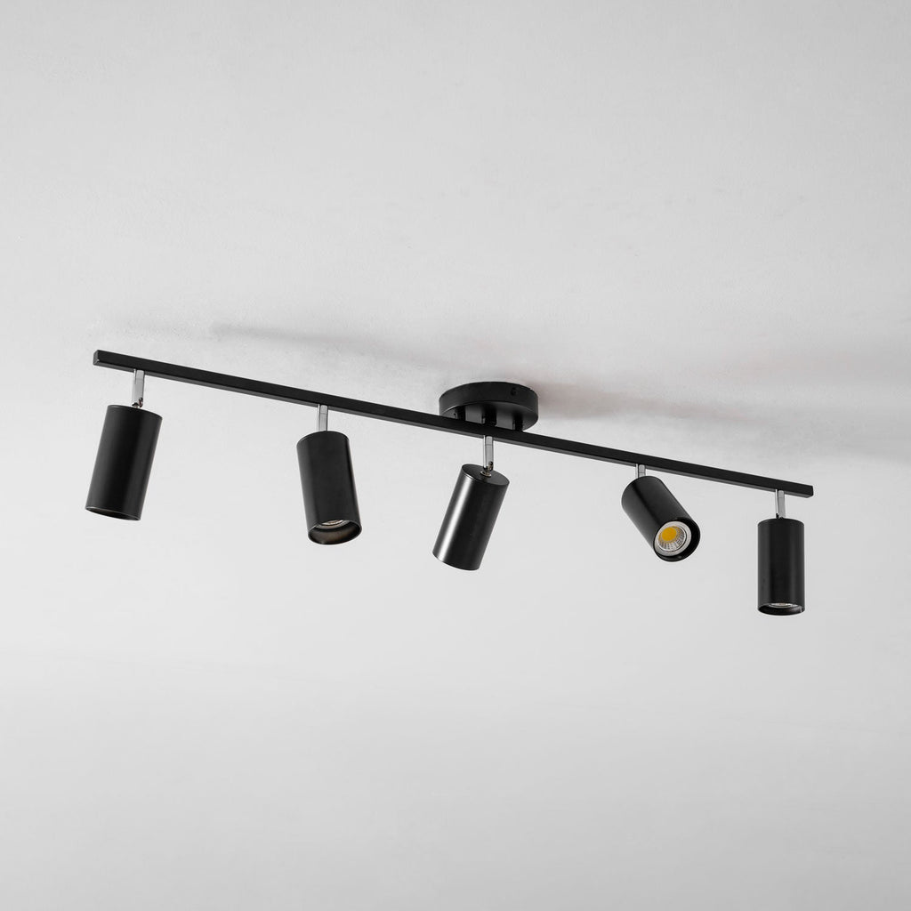 A black Clérachic five-head track light seen from an angle on a white ceiling, with one head glowing.