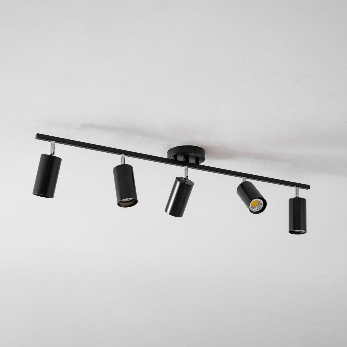 A black Clérachic five-head track light seen from an angle on a white ceiling, with one head glowing.