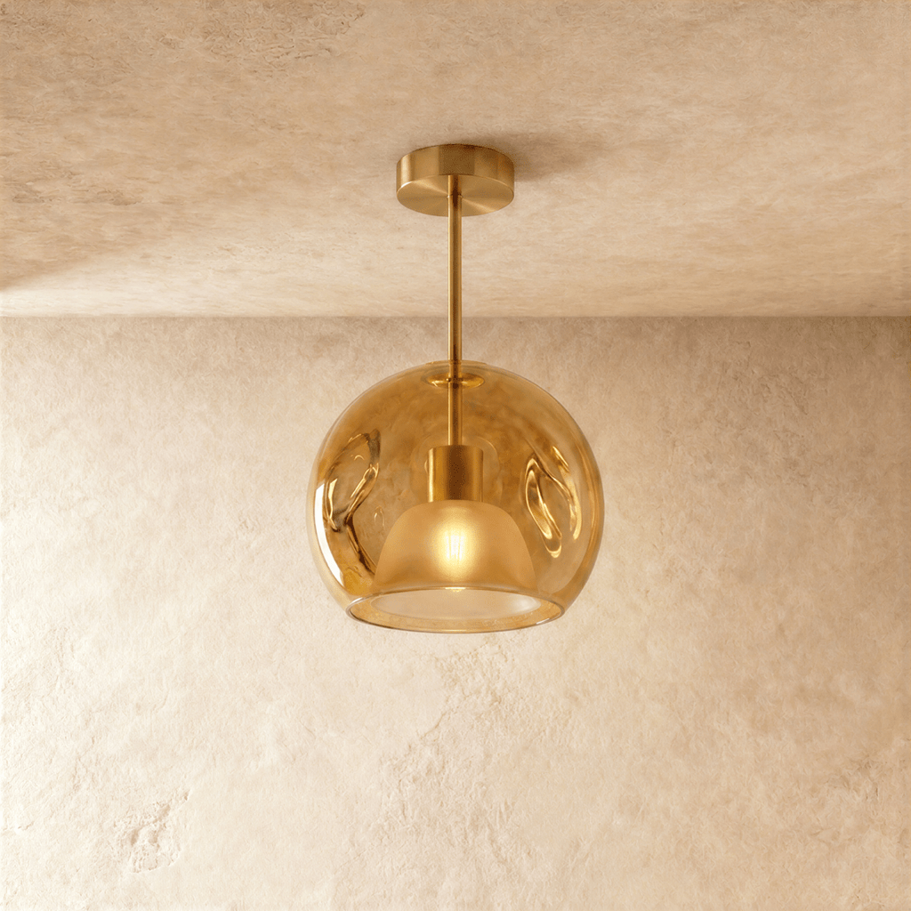Clérachic amber glass globe pendant drops on a gold rod from the ceiling, inner dome shade lit warm inside.