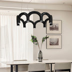 A black tiered-arch Clérachic chandelier above a white dining table, with framed prints and a vase of dark leaves behind.