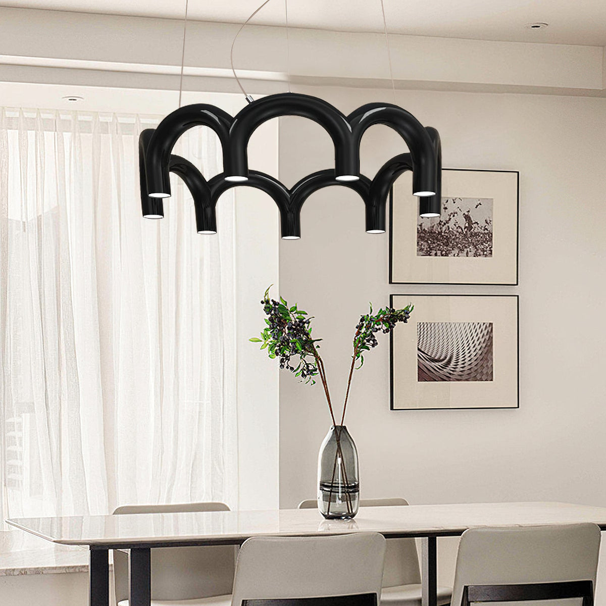 A black tiered-arch Clérachic chandelier above a white dining table, with framed prints and a vase of dark leaves behind.