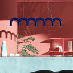 A blue arched-tube Clérachic chandelier in front of a coral wall with a monstera plant and a pale turquoise counter.