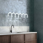A white arched-tube Clérachic chandelier against a rough grey concrete wall, above a kitchen counter with a black tap.
