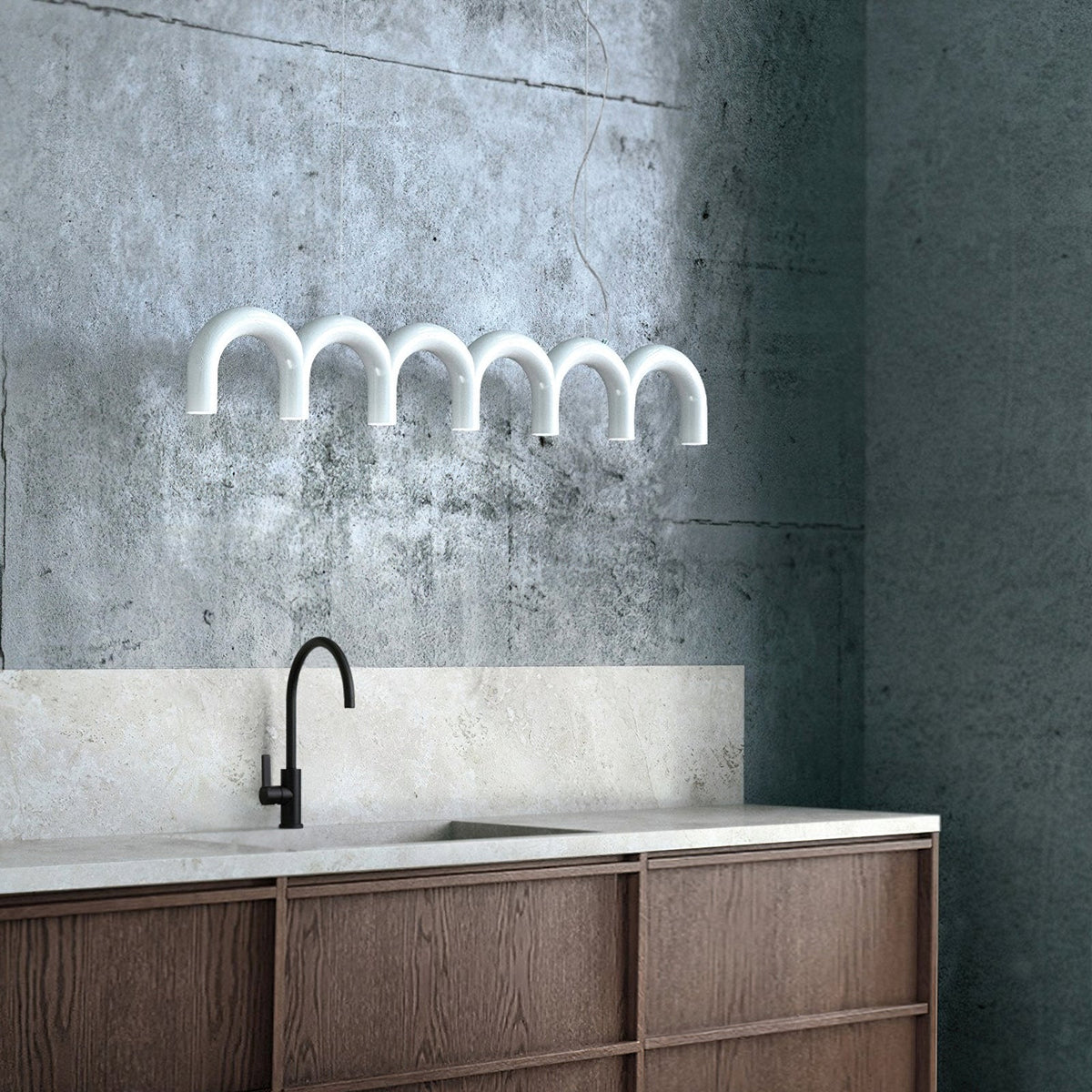 A white arched-tube Clérachic chandelier against a rough grey concrete wall, above a kitchen counter with a black tap.