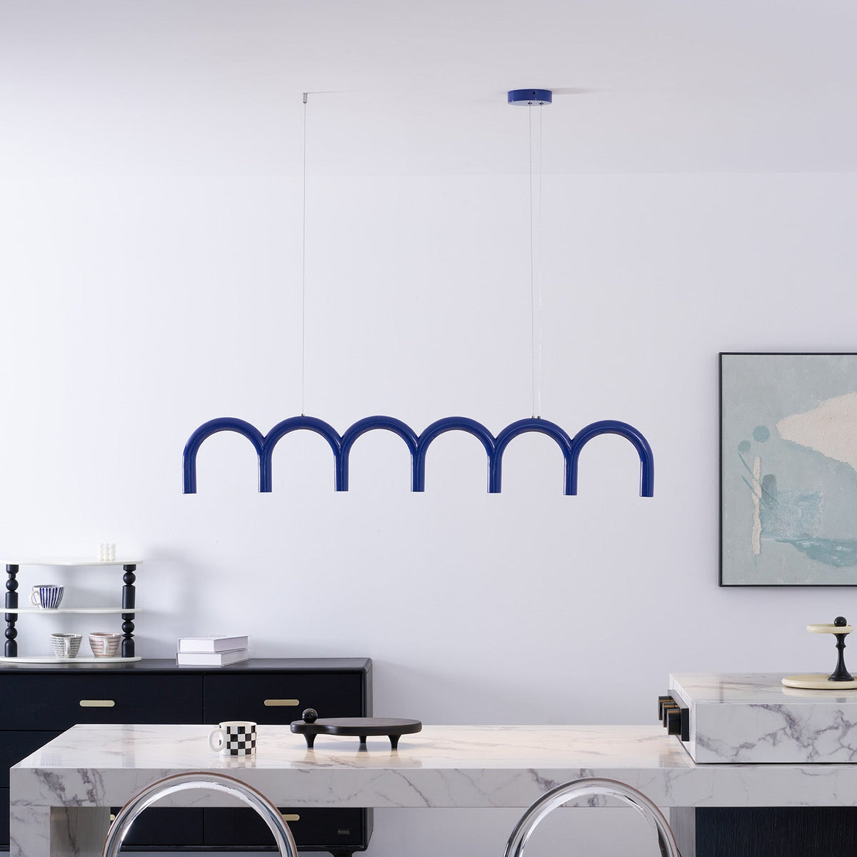 A blue arched Clérachic chandelier above a white marble counter, in front of a white wall and a framed print.