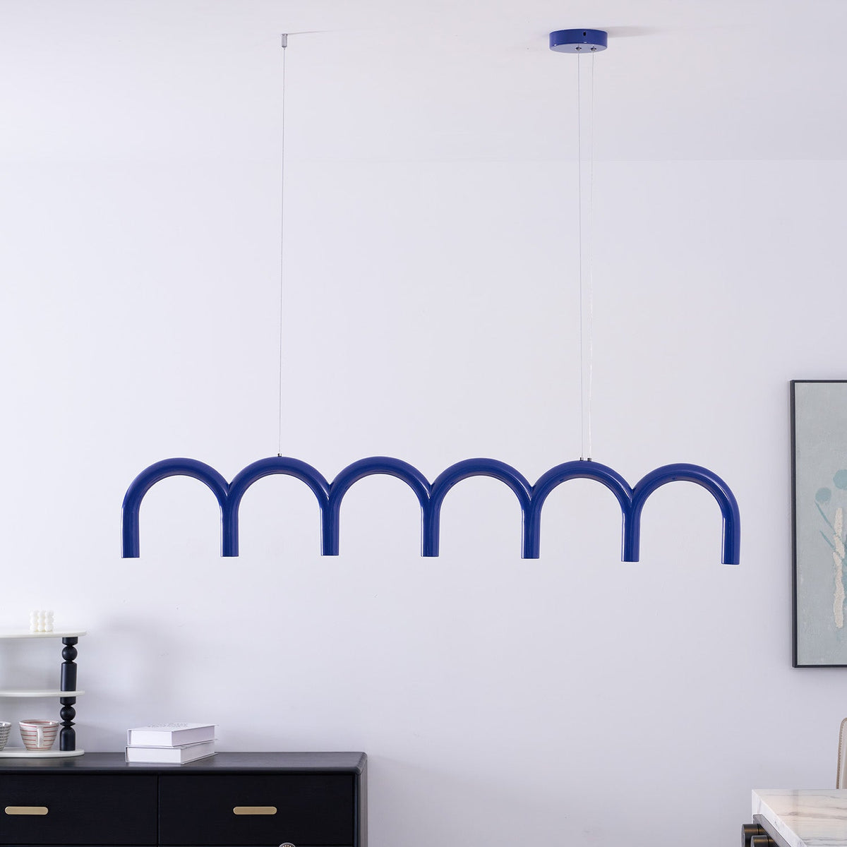 A blue arched Clérachic chandelier hung close to a white wall, with a framed print and a black sideboard behind.