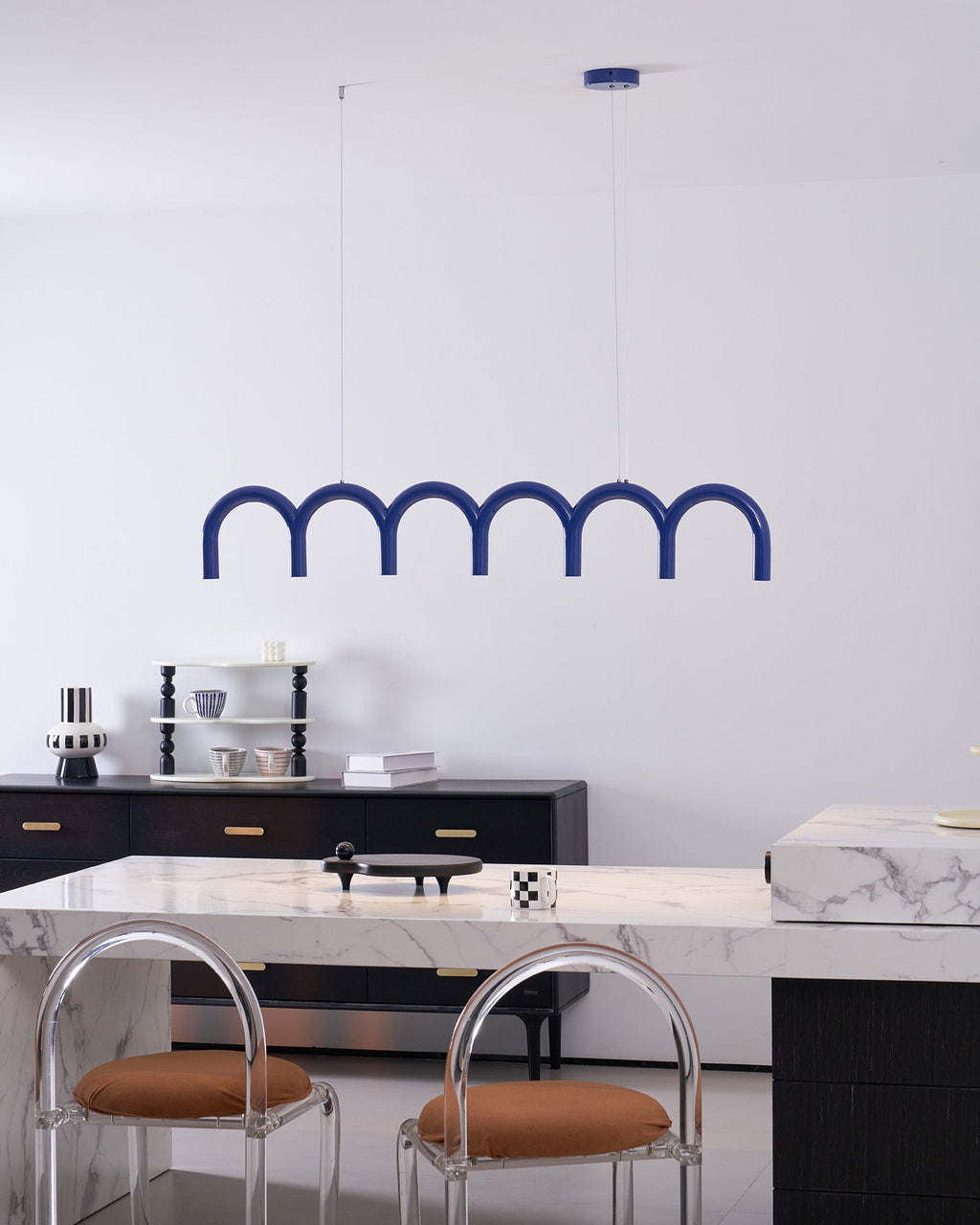 A blue arched Clérachic chandelier above a marble counter with two clear-backed stools with tan seats.