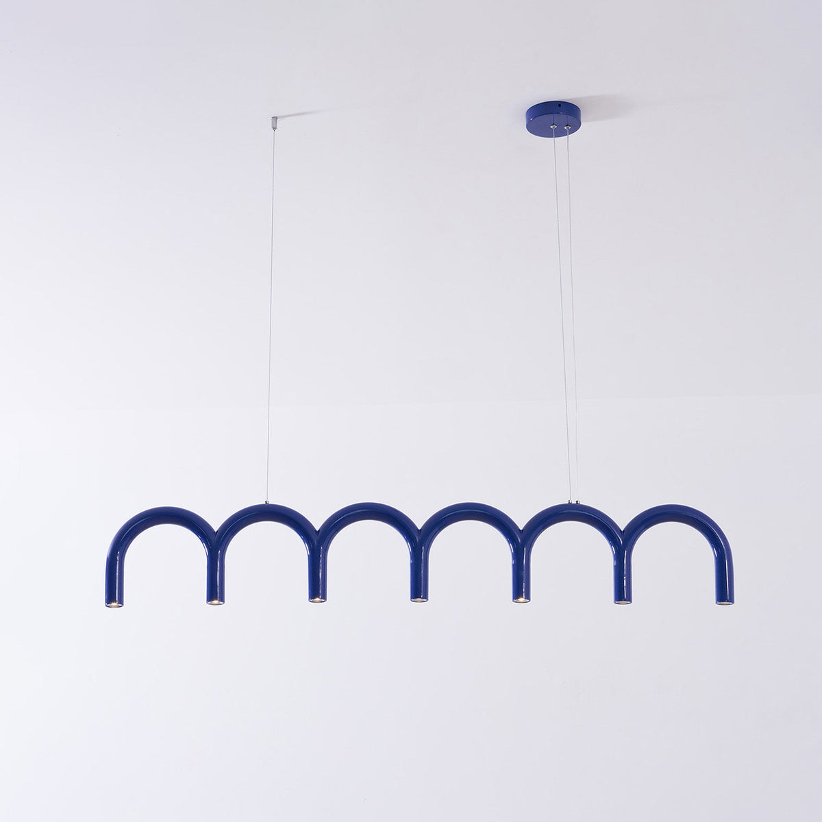 A blue arched Clérachic chandelier with a blue ceiling canopy and two thin cables, against a pale wall.