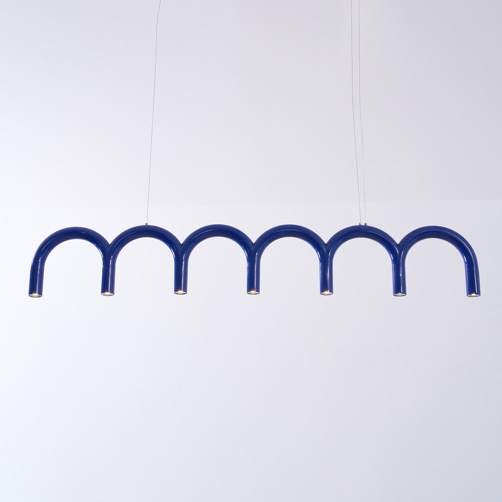 A long blue arched Clérachic chandelier hung on two thin cables, against a pale lilac backdrop.