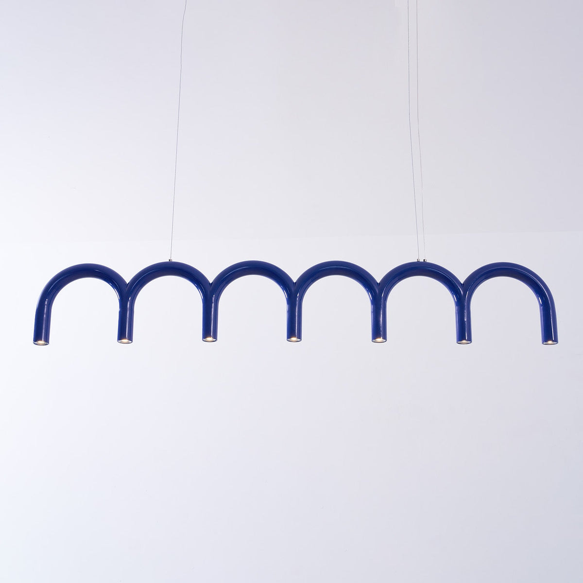 A long blue arched Clérachic chandelier hung on two thin cables, against a pale lilac backdrop.
