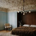 A Clérachic black Art Deco chandelier hangs above a bed in a bedroom with floral-painted ceiling and wallpaper.