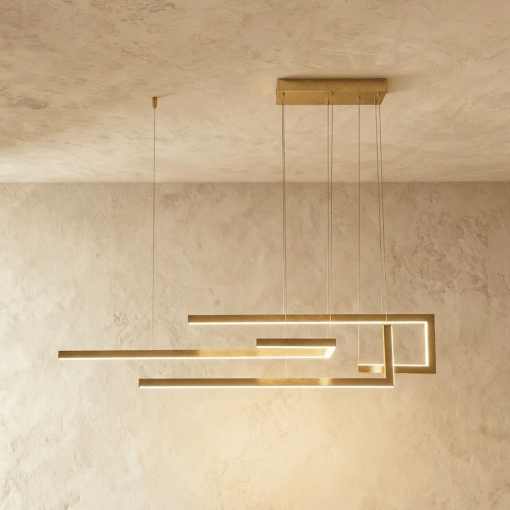 A side view of the Clérachic chandelier in gold, its geometric LED bars glowing warm against a plastered wall.