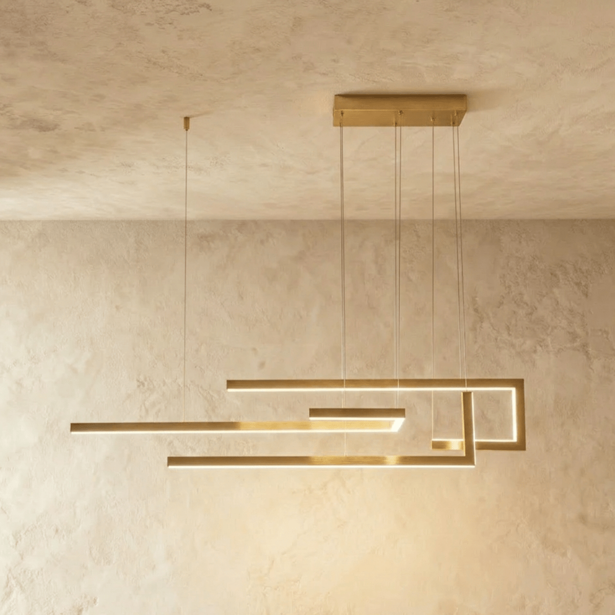 A side view of the Clérachic chandelier in gold, its geometric LED bars glowing warm against a plastered wall.
