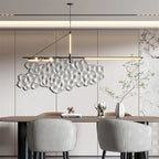 Clérachic black linear chandelier above a walnut dining table with grey pleated chairs, a sculptural wall panel and a vase of branches.