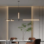 Clérachic black linear chandelier glowing warm above a marble dining table with rust-brown chairs and a leafy branch in a glass vase.