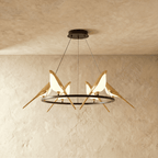 Clérachic ring chandelier with gold bird figures whose bodies glow white, hung from a black canopy in a plaster-toned studio.