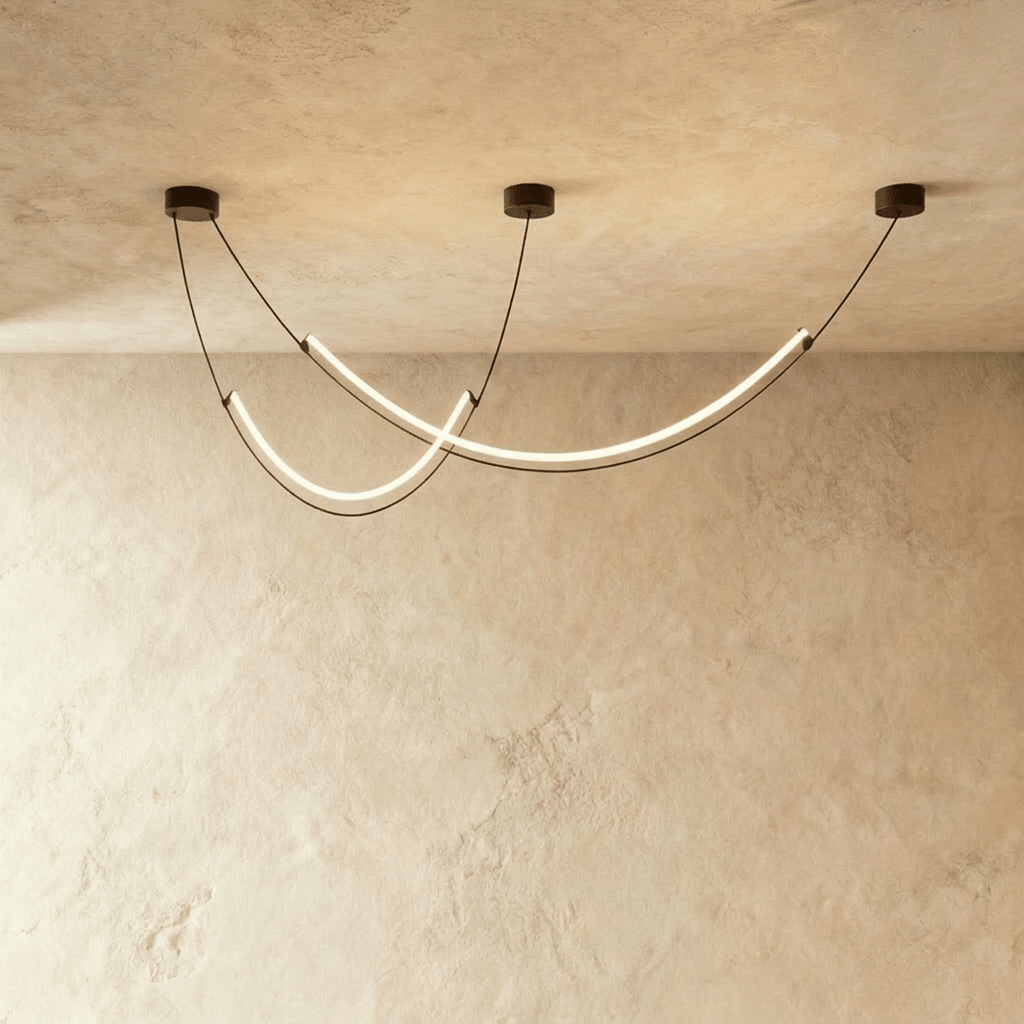 Clérachic chandelier of two crossing curved LED arcs on black cables, glowing warm white in a studio against limewash