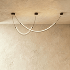 Clérachic chandelier of two crossing curved LED arcs on black cables, glowing warm white in a studio against limewash