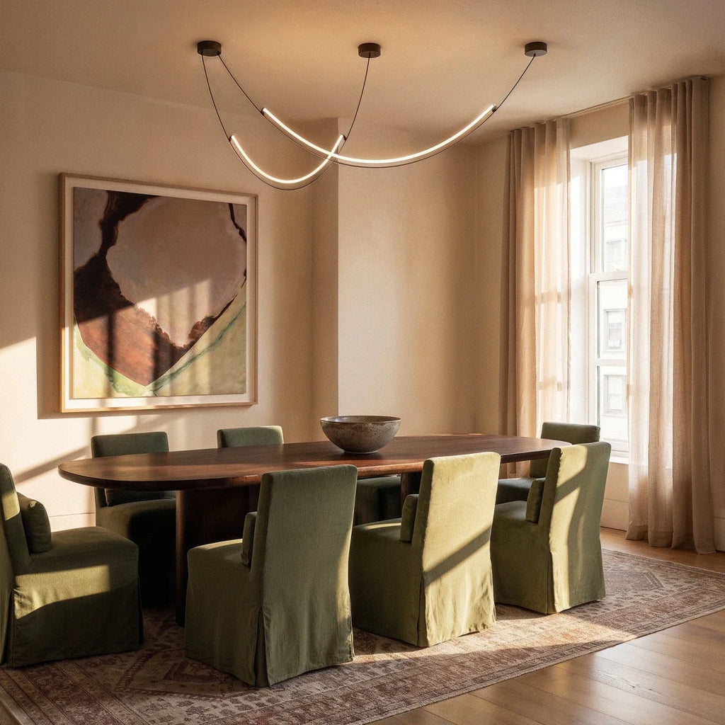 Clérachic curved LED arc chandelier lit above a long dining table with green chairs, a framed artwork and sheer curtains in the room