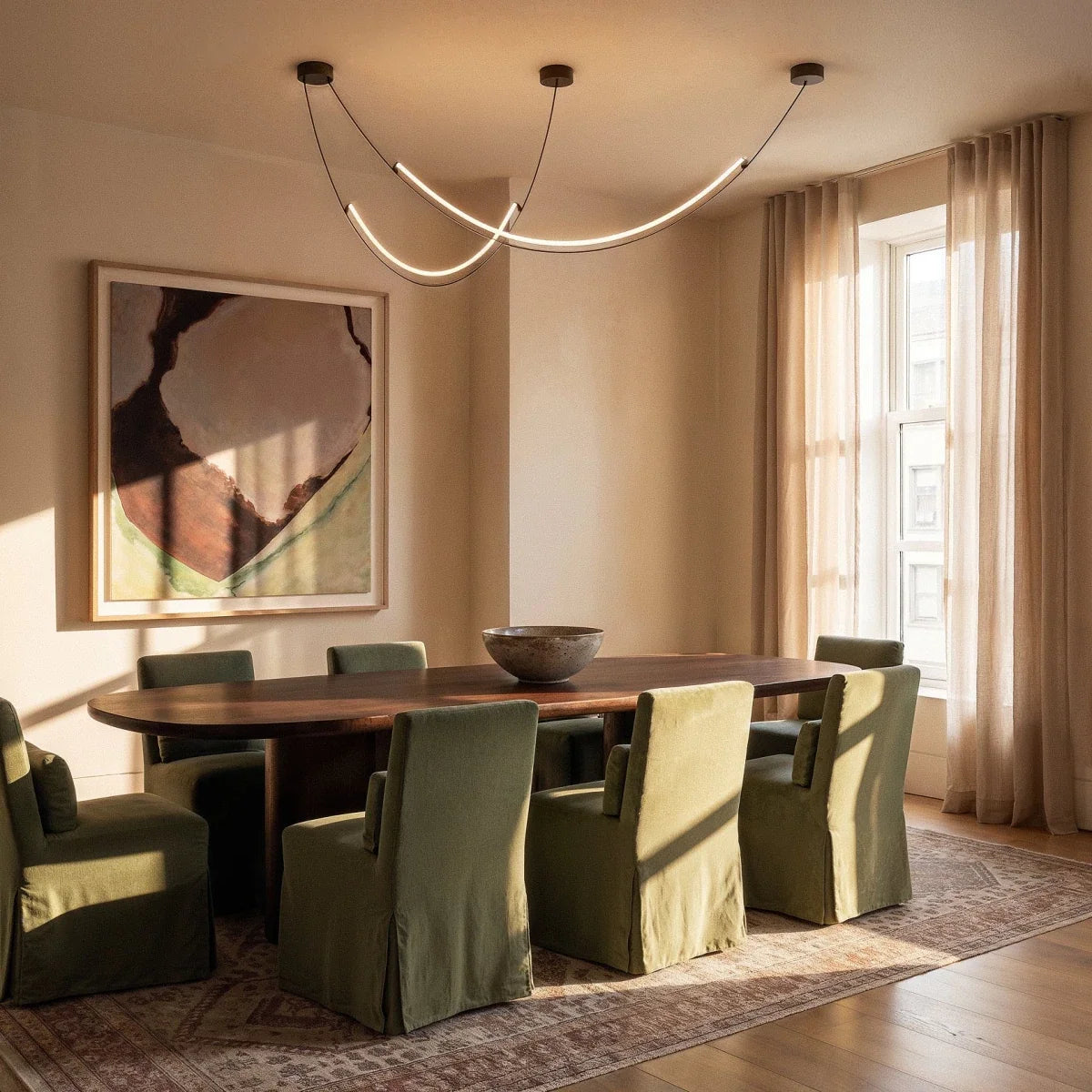 Clérachic curved LED arc chandelier lit above a long dining table with green chairs, a framed artwork and sheer curtains in the room