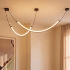 Clérachic chandelier of two crossing LED arcs on black cables lit in a warm room corner, beside sheer curtains and a framed print